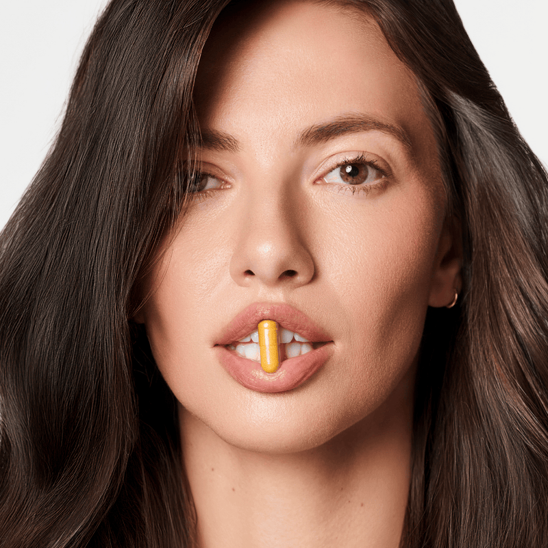 Woman with a yellow supplement capsule on her tongue against a neutral background