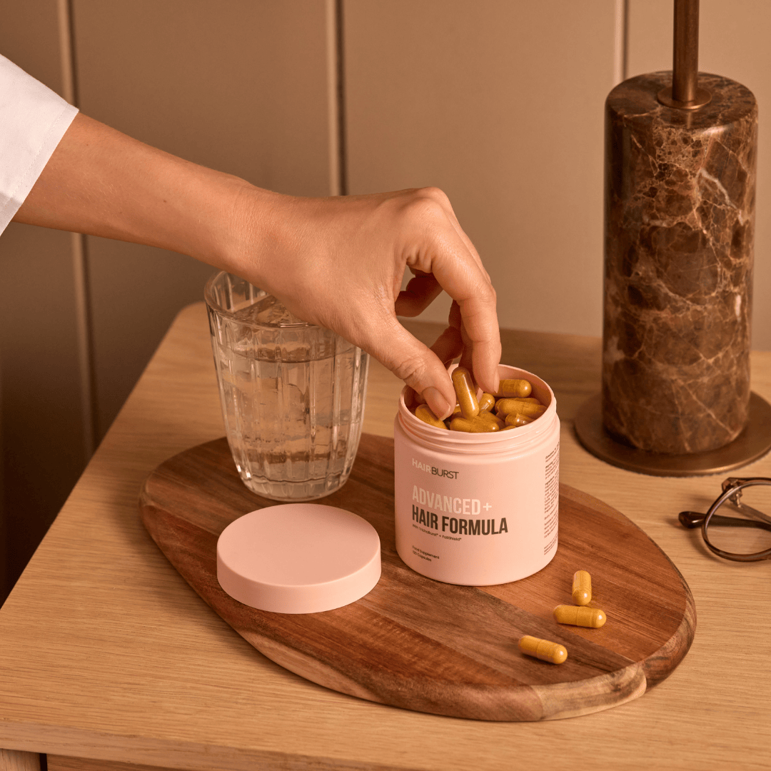 Person taking a supplement from a container labeled 'Advanced Hair Formula' on a wooden tray.